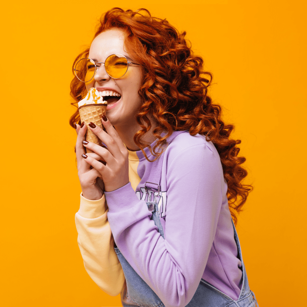 Young woman with red hair eats an ice cream cone from San Marco Dreamette on an orange background