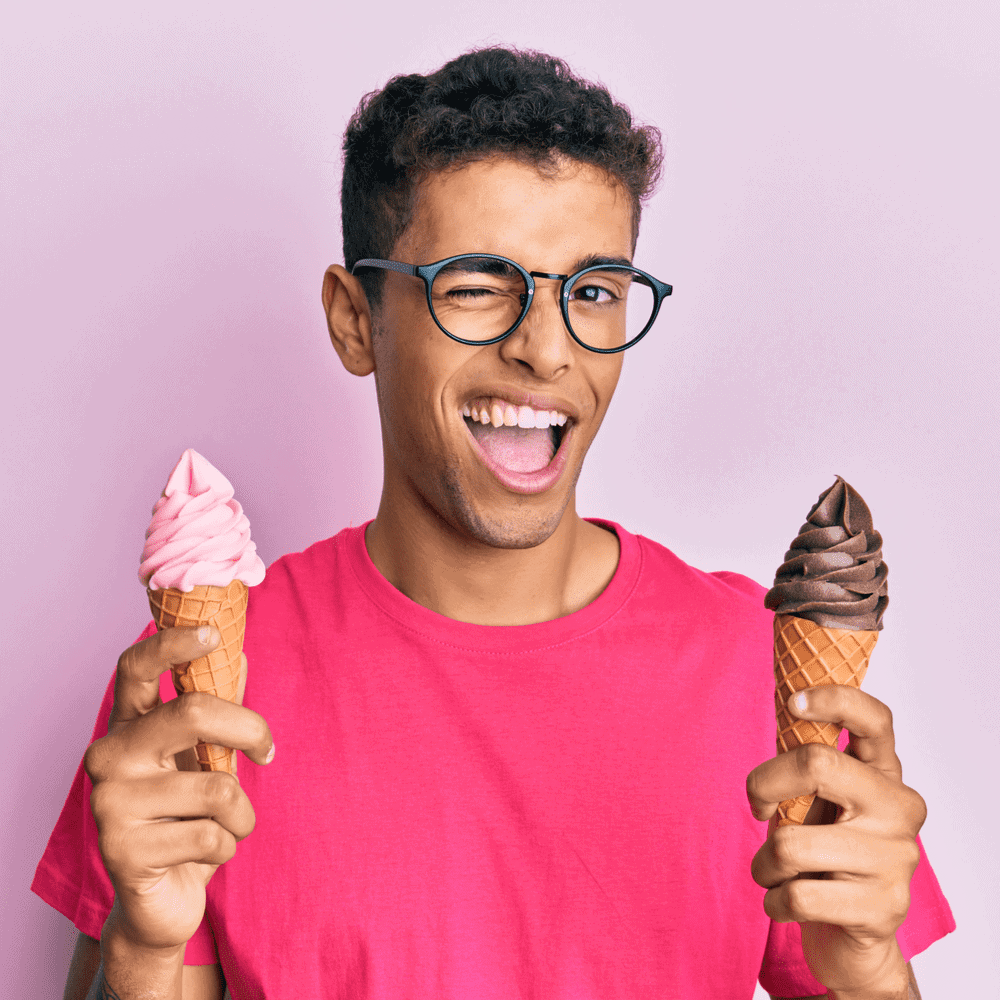 Young man holds two ice cream cones purchased from San Marco Dreamette on a pink background. We winks at the camera and smiles playfully.